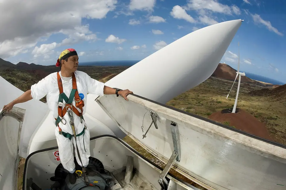 A technician in a safety harness stands on the hub of a wind turbine, one rotor blade above him.