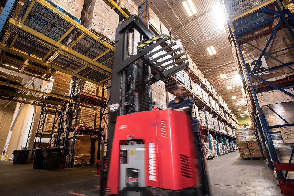 A red reach truck moving between tall pallet racks in a warehouse, its operator seated.